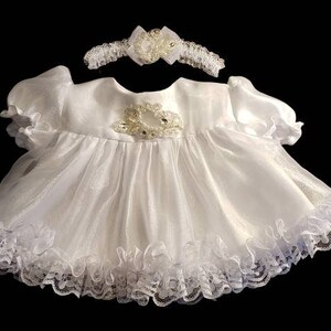 Peut inclure: Robe de bébé en satin blanc avec une bordure en dentelle et un embellissement perlé sur le corsage. La robe est accompagnée d'un bandeau en satin blanc assorti avec un embellissement perlé.