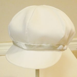 Pu&ograve; includere: Un cappello da marinaio bianco con una fascia in raso e due bottoni bianchi sul lato.