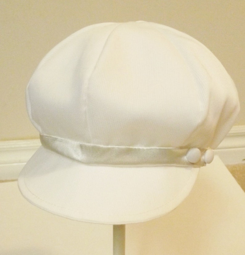 Pu&ograve; includere: Un cappello da marinaio bianco con una fascia in raso e due bottoni bianchi sul lato.