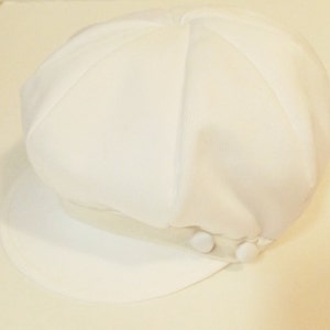 Pu&ograve; includere: Un cappello da newsboy in cotone bianco con una visiera bianca e due bottoni bianchi sul lato.