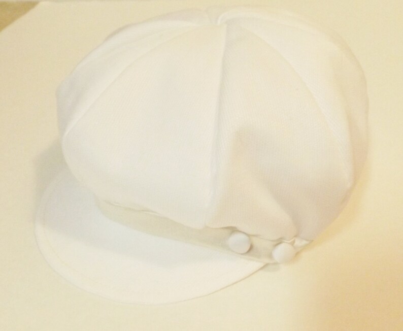 Pu&ograve; includere: Un cappello da newsboy in cotone bianco con una visiera bianca e due bottoni bianchi sul lato.
