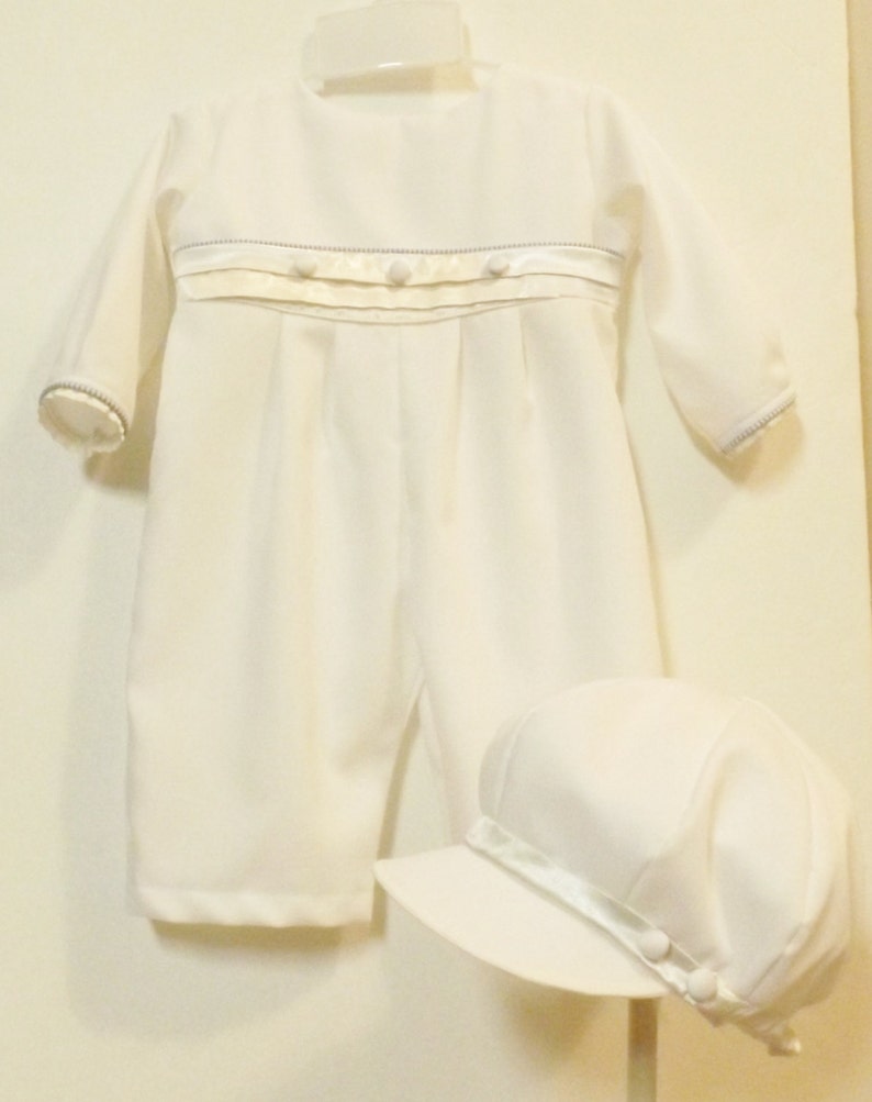 Pu&ograve; includere: Completo bianco per neonato con maniche lunghe e cappello bianco abbinato. Il completo &egrave; decorato con un nastro di raso bianco e bottoni perlati.