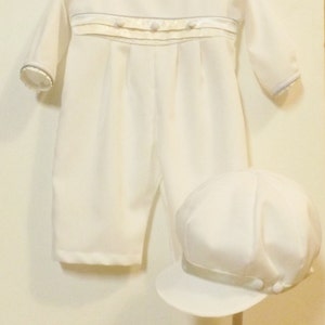Pu&ograve; includere: Completo bianco per neonato con maniche lunghe e cappello bianco abbinato. Il completo &egrave; decorato con un nastro di raso bianco con piccoli bottoni bianchi e un bordo blu intorno al collo e alle maniche. Il cappello ha un nastro di raso bianco e un piccolo bottone bianco.