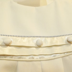 Pu&ograve; includere: Un vestitino bianco per neonata con un nastro di raso e tre bottoni bianchi. Il vestito ha un delicato bordo blu intorno al collo.