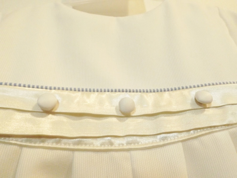Pu&ograve; includere: Un vestitino bianco per neonata con un nastro di raso e tre bottoni bianchi. Il vestito ha un delicato bordo blu intorno al collo.
