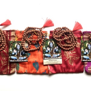 Puede incluir: Cuatro juegos de cuentas de mala Rudraksha marrones, cada uno con 108 cuentas, se muestran en sus bolsas individuales de cordón rojo, naranja, morado y rosa. Cada bolsa presenta un diseño de hojas doradas y una pequeña tarjeta con una imagen de Shiva y el texto "Mala de Rudraksha".
