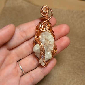 May include: A handcrafted copper wire pendant featuring a rough, off-white crystal. The crystal is intricately wrapped with copper wire, with decorative swirls and small, light pink beads. The pendant is designed to be worn as a necklace.