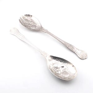 Puede incluir: Un par de utensilios de servicio de metal plateado que consta de una cuchara ranurada y una cuchara sólida. Ambas piezas presentan patrones en relieve de frutas y hojas en los cuencos y mangos, con un motivo de concha en el extremo.