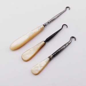 May include: Three antique crochet hooks with mother-of-pearl handles and metal hooks. The hooks vary in size and have a vintage aesthetic. The handles are a creamy white color, and the metal shows signs of age.