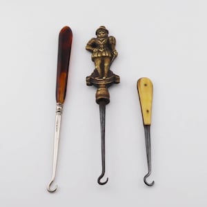 May include: Three antique tools with hook-shaped ends. One has a brown handle and silver shaft, another features a brass figure on top, and the third has a cream-colored handle. These tools are likely for sewing or crafting.
