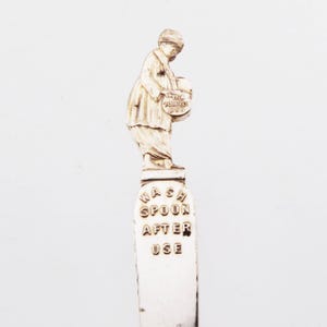 May include: A silver-toned metal spoon handle featuring a sculpted figure wearing a hat and long clothing, holding a small round object labeled THE TOM-TO PAN. Below the figure, the text WASH SPOON AFTER USE is embossed in capital letters.