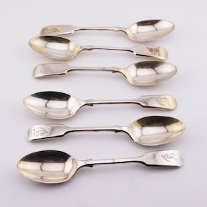 May include: A set of six silver-toned metal spoons arranged in rows. Each spoon features a polished oval bowl and a flat, squared handle end. Several of the handles are engraved with a decorative, swirling script letter monogram detail.