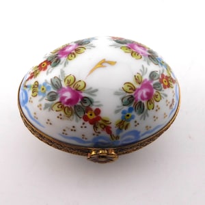 May include: An oval porcelain trinket box with a white base, decorated with hand-painted pink, yellow, and red floral clusters. The lid features a gold-coloured accent mark, and the base is encased in an ornate, textured brass metal rim with a flower-shaped clasp.