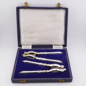 May include: A set of two silver-toned metal nutcrackers presented in a rectangular hinged storage case. The case features a deep purple velvet interior lining with moulded indentations for each tool and a white satin-lined lid interior.