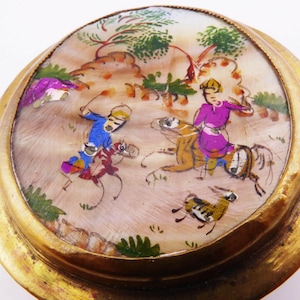 May include: A round, antique gold-toned trinket box with a painted scene of figures on horseback. The artwork features a landscape with trees and a running animal. The box has a detailed gold rim.