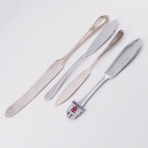 Può includere: Quattro coltelli da burro d'argento di vari design sono disposti su una superficie bianca. I coltelli hanno diverse forme di manico e stili di lama. Un coltello ha un manico decorativo con un emblema rosso e bianco.