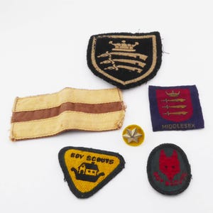 May include: A selection of vintage Boy Scout patches and badges. The patches display diverse designs, including a shield with a crown, a yellow triangle with a boat, and a red fox head. The badges are in various shapes and colours.