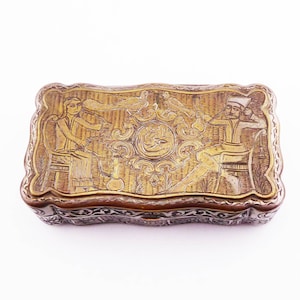 May include: A rectangular brass box featuring an intricate engraved scene on the lid. The design depicts two figures seated on chairs, separated by a central circular medallion containing Arabic script, with birds and floral motifs surrounding them.
