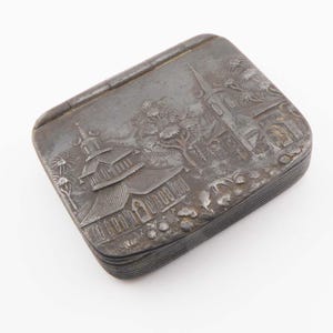 May include: A rectangular, antique silver-coloured box with a hinged lid. The lid features a detailed relief scene of buildings, trees, and foliage. The box has a slightly worn appearance, suggesting age and use.
