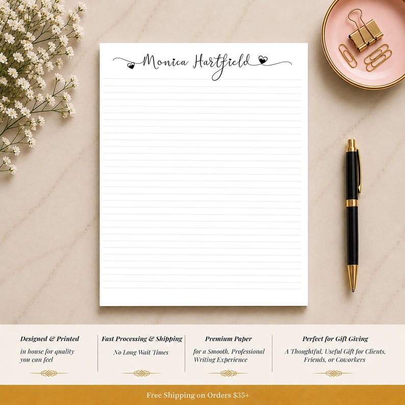 Op de afbeelding: Een wit gelijnd notitieblok met de naam "Monica Hartfield" in elegant schrift. Een zwarte pen met gouden accenten en een roze schaal met gouden paperclips zijn ook zichtbaar. Het notitieblok is ontworpen en gedrukt voor kwaliteit.