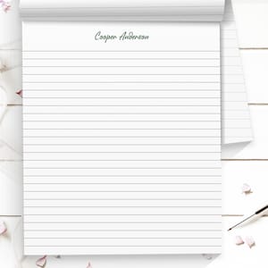 May include: A white notepad with horizontal gray lines and the text "Cooper Anderson" in green script. The notepad is partially rolled up, revealing more lined pages. A pen and flower petals are in the background.