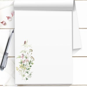 May include: A white notepad with a floral watercolor design. The notepad is open, revealing blank pages. A silver pen with a crystal-like design rests on the left. The notepad is on a white wooden surface.