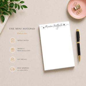 Op de afbeelding: Een witte mini-notitieblok met de tekst "Monica Hartfield" in elegant schrift. Een zwarte en gouden pen, een roze schaal met paperclips en een takje groen zijn ook aanwezig. Het notitieblok is ontworpen voor snelle notities, praktische cadeaus en onderweg.