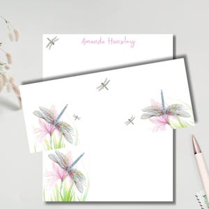 May include: White stationery set featuring watercolor illustrations of dragonflies and pink flowers. The top sheet has the name "Amanda Hansley" in pink script. The set includes a notepad and a larger sheet of paper.
