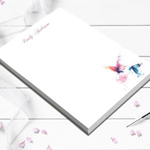 May include: White notepad with the name "Emily Anderson" in cursive script. Watercolor butterflies in shades of pink, orange, and blue are in the corner. A black pen and pink flower petals are nearby.