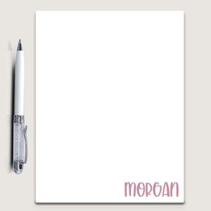 May include: A white notepad with the name "MORGAN" in pink script at the bottom right. A white pen with a crystal-like barrel is positioned to the left of the notepad. The notepad and pen are on a light gray surface.