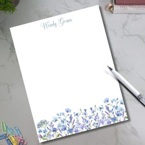 May include: White stationery with a floral border in shades of blue and purple. The name "Wendy Gram" is written in script at the top. A pen rests on the side, ready for use. Stationery for writing letters.