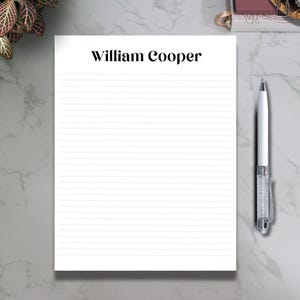 May include: A white lined notepad with the name "William Cooper" printed at the top. A silver pen with a crystal-like design rests on a marble surface, next to the notepad. A plant and paper clip are also visible.