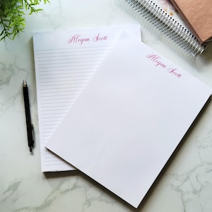 May include: Two white personalized notepads with the name "Megan Scott" printed in pink script. One notepad has lined pages, the other is blank. A black pen and a spiral notebook are also visible.