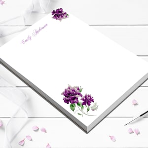 May include: White notepad with purple floral accents and the name "Emily Anderson" printed in purple script. The notepad features a stack of white pages and a dark gray edge. A pen and scattered pink flower petals are also visible.