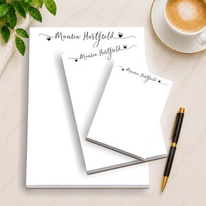 Op de afbeelding: Een set van drie witte notitieblokken van verschillende formaten, elk met de naam "Monica Hartfield" in elegant zwart schrift. Het ontwerp bevat kleine hartaccenten. Een zwarte en gouden pen ligt in de buurt, met een kopje koffie op de achtergrond.