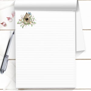 May include: A white notepad with horizontal lines and a decorative birdhouse illustration. The birdhouse is brown with a heart-shaped opening, surrounded by green leaves and small birds. A silver pen rests on the left side.