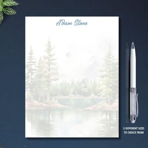 May include: A notepad with a scenic landscape design featuring a lake, trees, and mountains. The name "Adam Stone" is printed at the top. A white pen with a silver clip and a decorative barrel is placed next to the notepad. Text at the bottom reads "5 DIFFERENT SIZES TO CHOICE FROM".