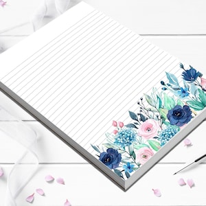 May include: A white notepad with lined pages and a floral design. The floral arrangement features blue, pink, and teal flowers and leaves. The notepad is rectangular and sits on a white surface.