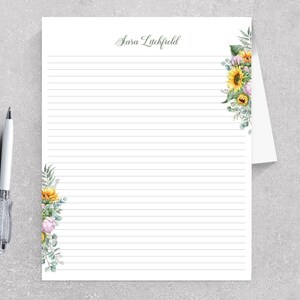 May include: White lined stationery with floral accents and the name "Sara Litchfield" in script. The stationery features a watercolor design of sunflowers, greenery, and other flowers. A silver and white pen rests on the side.