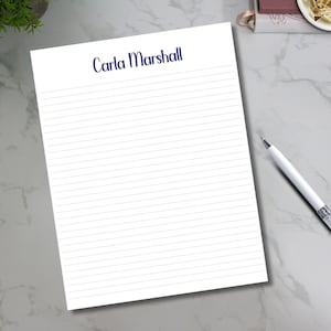 May include: A white lined notepad with the name "Carla Marshall" in navy blue script at the top. A white pen with a silver clip rests on a gray surface. Colorful paper clips are in the lower left corner.