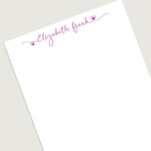 May include: A white notepad with the name "Elizabeth Frank" in a pink, cursive font. Two small pink hearts are on either side of the name, adding a touch of elegance to the stationery.