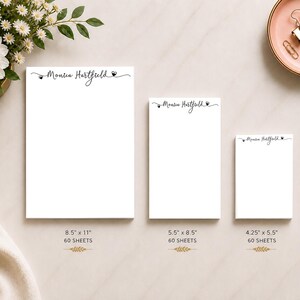 Op de afbeelding: Drie witte notitieblokken van verschillende formaten, elk met de naam "Monica Hartfield" in sierlijke letters. Het grootste notitieblok is 21,6 cm x 27,9 cm met 60 vellen. Een gouden pen en een roze schaal met paperclips zijn ook zichtbaar.