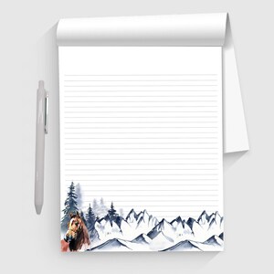 May include: A lined notepad with a watercolor design of a horse, pine trees, and mountains in shades of blue and brown. A silver pen rests on the left side of the notepad.