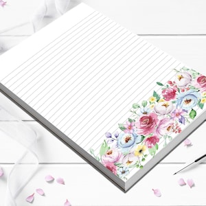 May include: A white lined notepad with a floral design. The floral design features pink, blue, and yellow flowers. The notepad is on a white wooden surface with pink flower petals scattered around.
