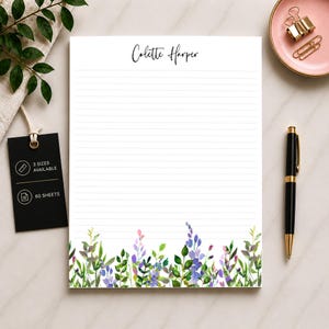 May include: A lined notepad with the name "Colette Harper" in cursive script at the top. The bottom edge features a watercolor floral design in shades of green, purple, and pink. A black pen and pink dish with gold paper clips are on the right.