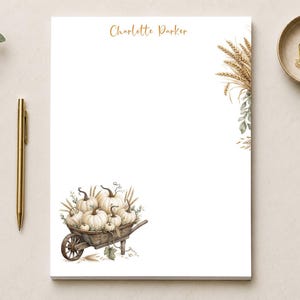 May include: A white rectangular notepad featuring the name Charlotte Parker in gold script at the top. The bottom left corner displays a watercolor illustration of a wooden wheelbarrow filled with white pumpkins, while wheat stalks decorate the right side.