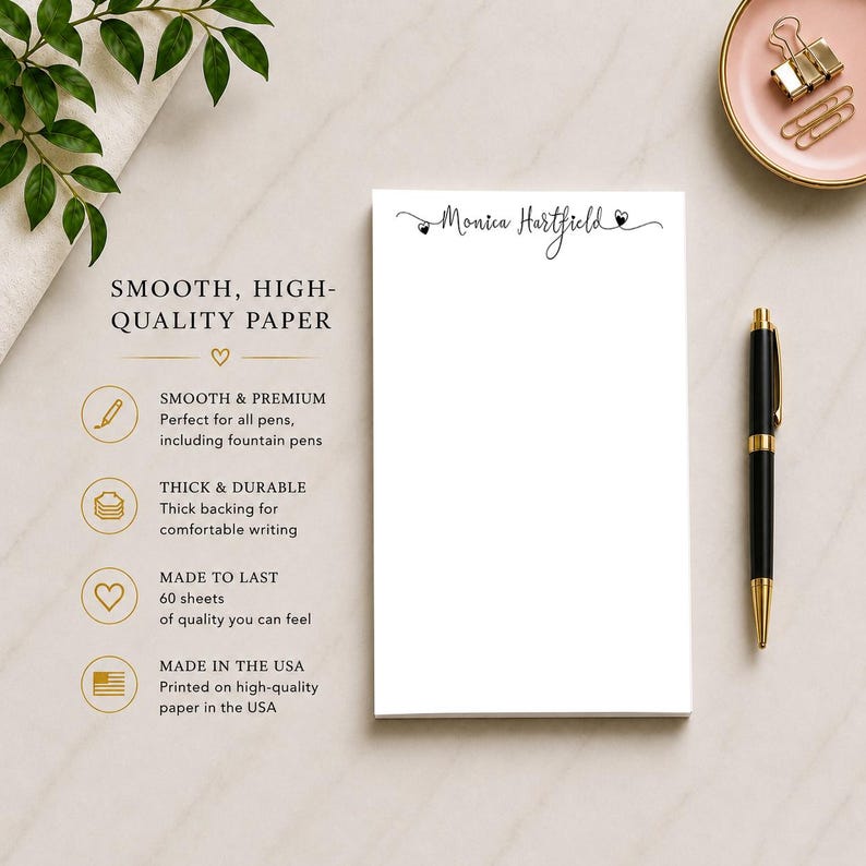 Op de afbeelding: Een witte notitieblok met de naam "Monica Hartfield" in script. Een zwarte en gouden pen en een klein roze schaaltje met paperclips liggen naast het notitieblok. Tekst op het notitieblok luidt "Smooth, High-Quality Paper".