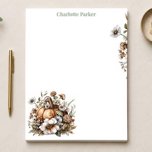 May include: A white rectangular notepad featuring the printed name Charlotte Parker at the top center. The bottom left corner displays a detailed illustration of an orange pumpkin surrounded by white and brown flowers, leaves, and greenery.
