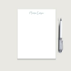May include: A white notepad with the name "Monroe Carson" in light blue script. A white pen with a crystal-like barrel and silver accents rests on the right side of the notepad. The background is a light gray.