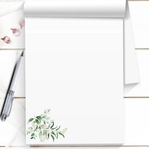 May include: A white notepad with a floral design in the corner, featuring green leaves and small white flowers. A silver pen rests on the side, with a few pink petals scattered nearby. The notepad is on a white wooden surface.